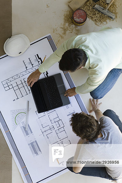 Two men crouching looking over blueprints  using laptop set on floor
