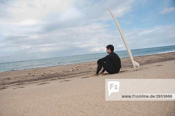 Junger Mann sitzt am Strand mit seinem Surfbrett