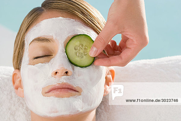 Young woman wearing facial mask having cucumber slice placed over eye