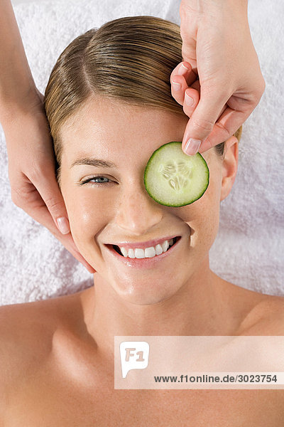 Smiling young woman lying on massage table with cucumber slice being placed over eye