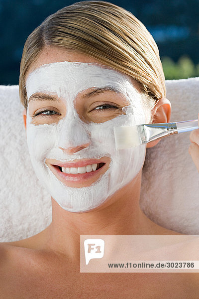 Lächelnde junge Frau bürstet Gesichtsmaske auf Gesicht