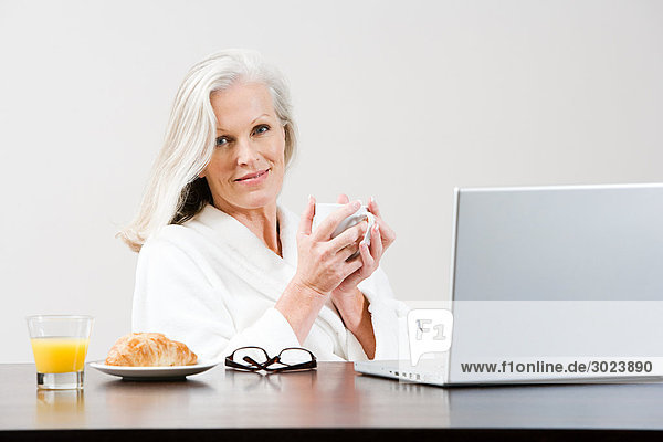 Middle aged woman eating breakfast while using laptop