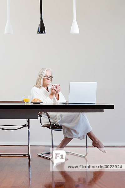 Middle aged woman sitting at table with breakfast and laptop