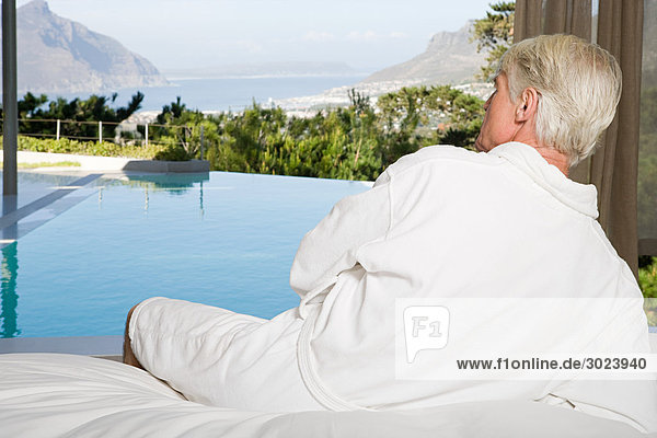 Middle aged man wearing bathrobe lying on bed and looking out at swimming pool
