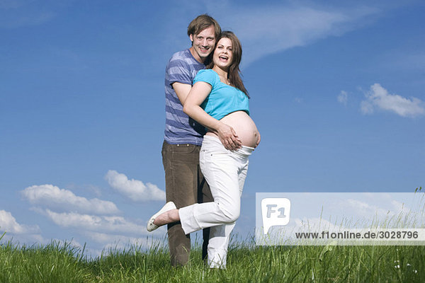 Junger Mann umarmt schwangere Frau  auf der Wiese stehend  lächelnd  Portrait