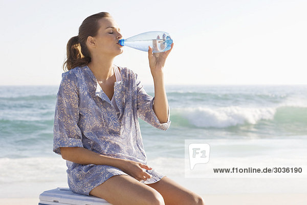 Frau sitzt auf einer Eisbox und trinkt Wasser am Strand.