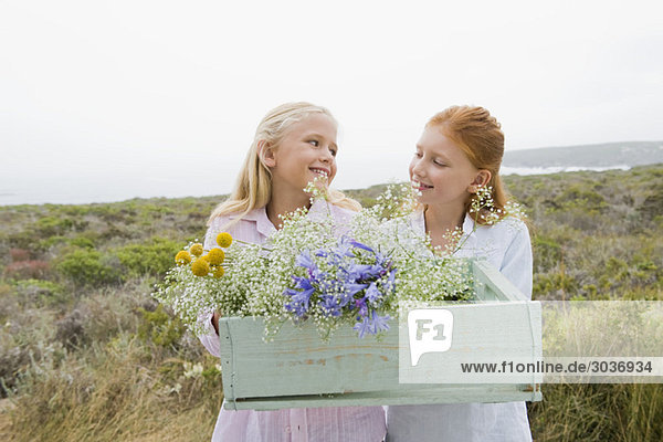 Zwei Mädchen  die eine Schachtel mit Blumen tragen und lächeln.