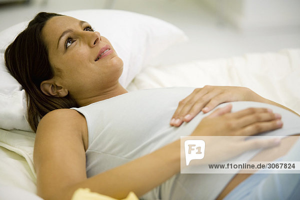 Schwangere Frau auf dem Rücken liegend mit Händen auf dem Bauch  aufblickend und lächelnd