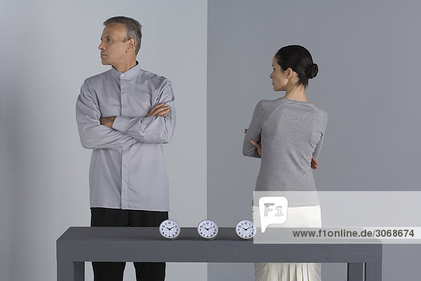 Couple standing with arms folded  backs turned to each other  clocks on table in front of them