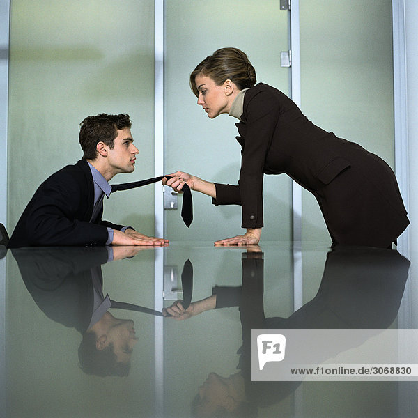 Mann und Frau von Angesicht zu Angesicht am Tisch  Frau lehnt sich nach vorne und zieht die Krawatte des Mannes.