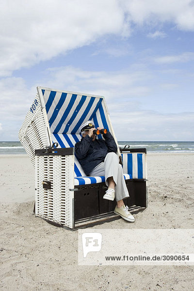 Ein älterer Mann sitzt in einem Strandkorb mit Kapuze und schaut durch ein Fernglas.