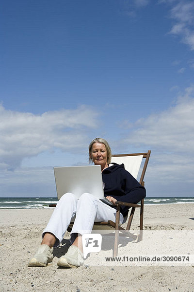 A senior woman sitting in a lounge chair at the beach using a laptop