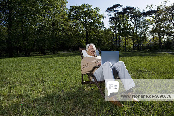 Ein älterer Mann  der in einem Sessel sitzt und einen Laptop benutzt  im Freien.