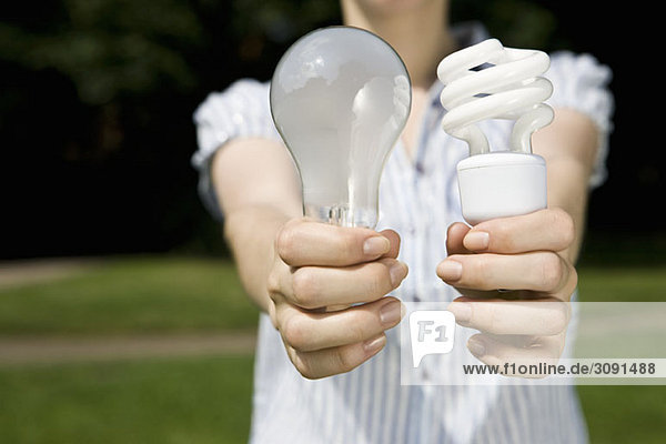 Eine junge Frau hält eine normale Glühbirne und eine Energiesparlampe in der Hand.