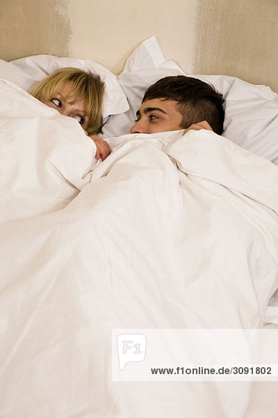 A young couple in bed with the duvet pulled up to their eyes