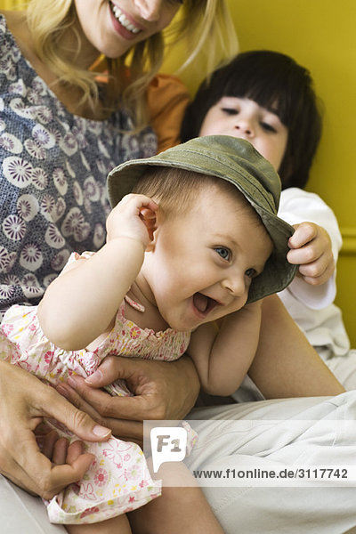 Kleinkind sitzend mit Mutter und Bruder  Hut tragend und lachend