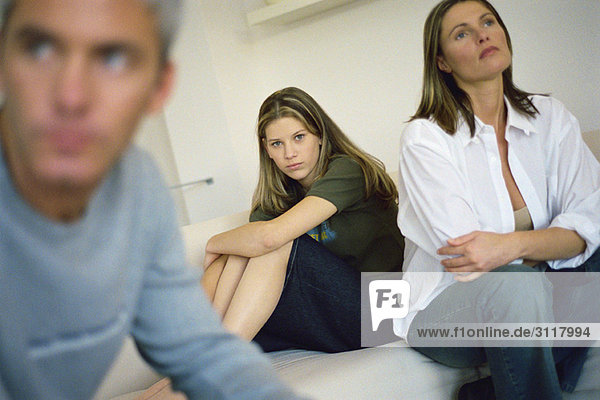 Teenager-Mädchen sitzt auf dem Bett und umarmt die Knie  Eltern sitzen in der Nähe und schauen mit frustrierter Luft weg.
