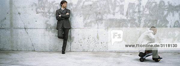 Businessman with arms folded leaning against wall watching associate ride child's tricycle