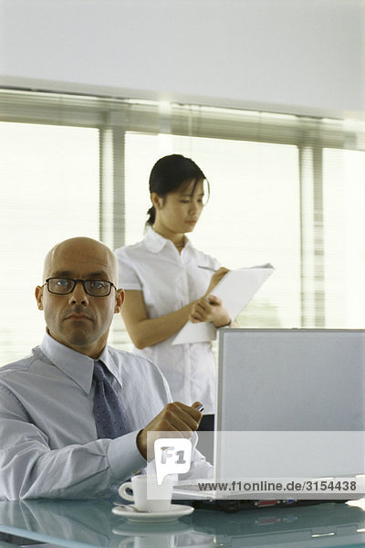 Businessman sitting at desk  looking at camera  female assistant taking notes in background