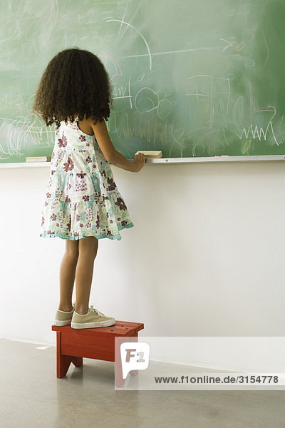 Little girl standing on stool in front of blackboard  rear view