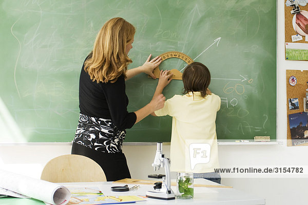 Teacher helping boy draw angle on blackboard using protractor  rear view