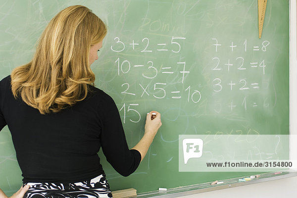 Elementary teacher writing arithmetic on blackboard  rear view