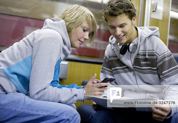 Two teenagers using mobile phone and print media in city train  slanted view