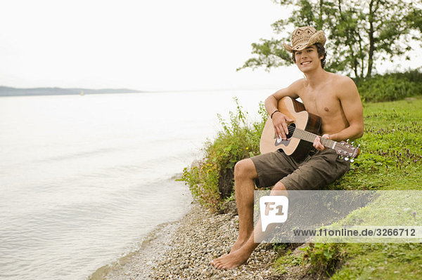 Starnberger See  Young man sits on lakeshore playing guitar  smiling  portrait