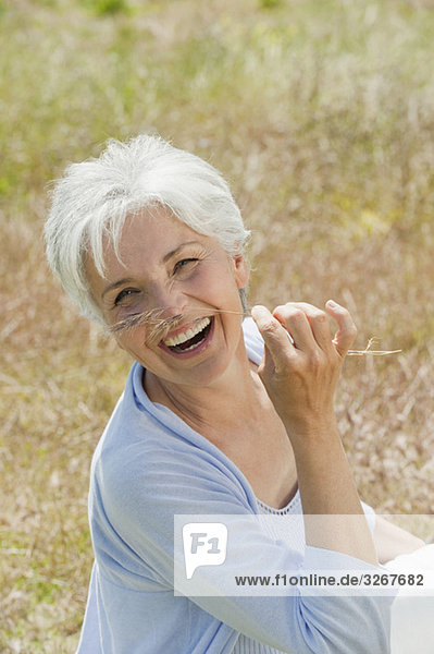 Spain  Mallorca  Senior woman sitting in meadow  holding blade of grass  laughing  elevated view