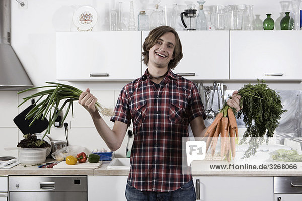 Junger Mann in der Küche mit Karotten und Frühlingszwiebeln  lächelnd  Portrait