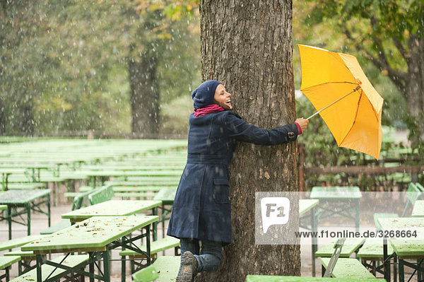 Englischer Garten  Frau im Biergarten umarmt Baum  hält Regenschirm  lachend