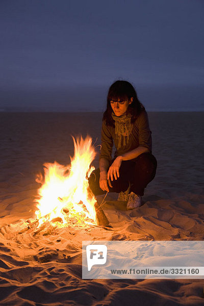 Eine Frau  die in ein Feuer starrt  das am Strand brennt.
