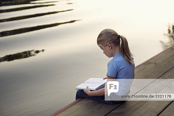 A young girl sitting at the edge of a jetty and reading a book