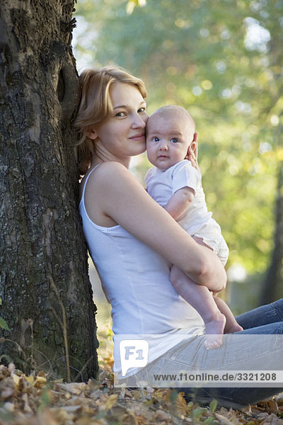 Eine Mutter sitzt an einem Baum  küsst ihr Baby und schaut in die Kamera.