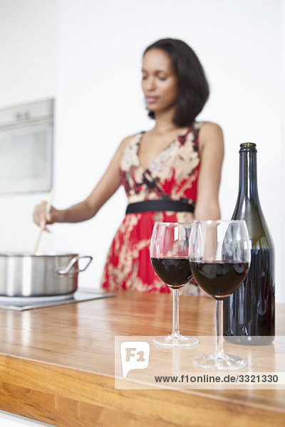 Zwei Gläser Rotwein und eine Weinflasche mit einer Frau  die im Hintergrund einen Topf rührt.