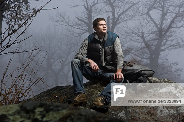 Ein männlicher Wanderer sitzt auf einem Felsen in einem nebligen Wald.