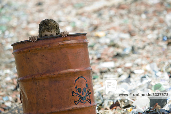 Kind schaut über den Rand eines mit Schädel und Knochen markierten Metallfasses  umgeben von einer Mülldeponie.