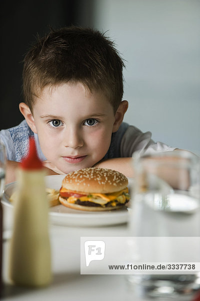 Kleiner Junge am Tisch sitzend mit Cheeseburger auf Teller  Kopf auf Armen liegend