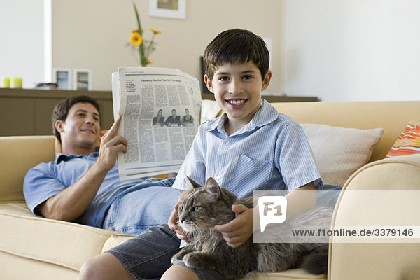 Young boy with pet cat on lap  father reading newspaper in background