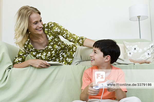 Kleiner Junge sitzt auf dem Boden und spielt ein Videospiel und schaut auf die Mutter  die sich auf dem Sofa entspannt.