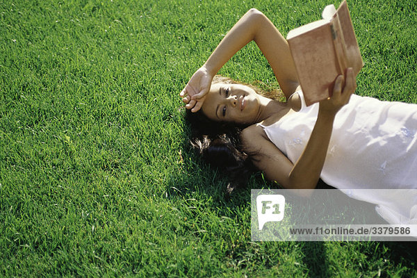 Junge Frau auf Gras liegend  Buch haltend  Kamera lächelnd