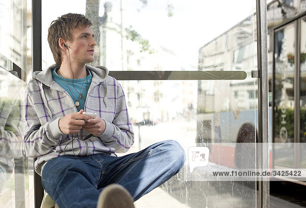 Junger Mann mit MP3-Player sitzt in einer Bushaltestelle  Flachwinkelansicht