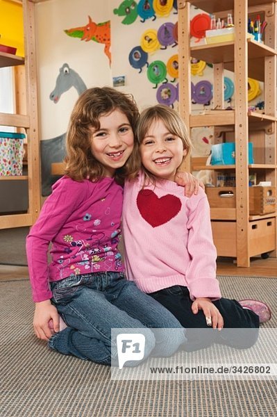 Deutschland  Zwei Mädchen (4-5)  (6-7) im Kinderzimmer auf dem Boden sitzend  umarmend  Portrait
