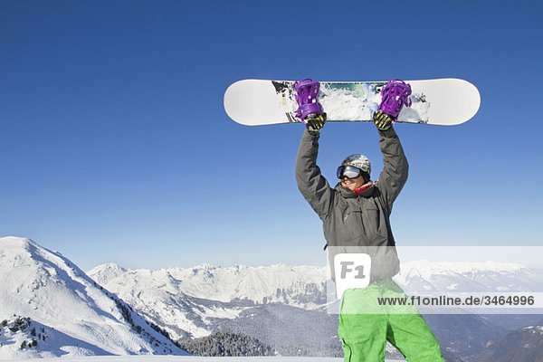 Junger Mann hält sein Snowboard über dem Kopf.