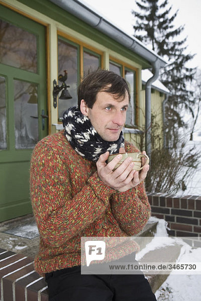 A man sitting on the front stoop of a house holding a hot drink