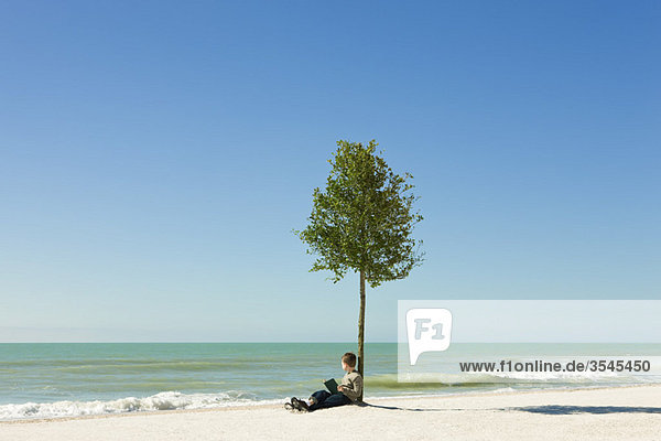Junge sitzt unter einem Baum am Strand mit einem Buch in der Hand und schaut auf das Meer.