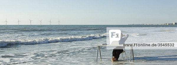 Mann entspannt am Schreibtisch am Strand  Windkraftanlagen am Horizont