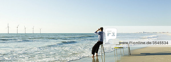 Mann auf Schreibtischkante am Wasserrand am Strand mit Blick auf Offshore-Windkraftanlagen am Horizont