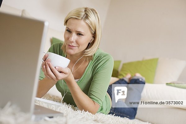 Junge Frau liegt auf einem Teppich mit Kaffeetasse  Ohrstöpseln und Laptop