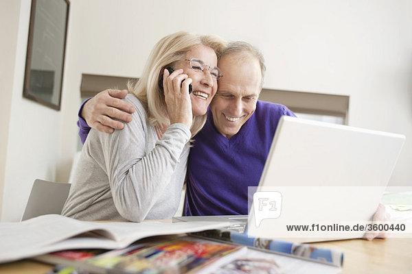 Frau  die mit ihrem Mann auf einem Handy vor einem Laptop sitzt.